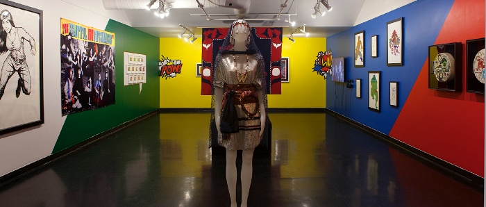 画廊的照片显示，在红黑相间的纽扣毯前，前景中穿着王冠的人体模特。画廊的墙壁被漆成明亮的颜色，顺时针环绕房间：白色、绿色、黄色、红色和蓝色。后墙的左右角都有漫画风格的图形，上面写着POW和WOW。墙上的艺术品从左到右依次为：一幅动作中的女超级英雄的黑白大图，一幅月亮般风景中几位土著妇女的黑白大旗，一个有13张小框架交易卡的喜剧演讲泡泡，一个视频显示器，6张黑框图纸，2张黑框彩绘隐藏鼓。