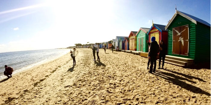 人们站在海滩上看水，身后有五颜六色的海滩盒子